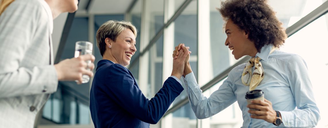 happy-businesswomen-holding-hands-while-greeting-hallway-1140x445